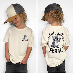 A young boy wearing a white t-shirt with a graphic of a raccoon and the text "CUTE BUT FERAL" printed on it.