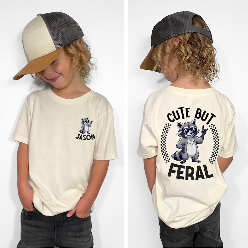 A young boy wearing a white t-shirt with a graphic of a raccoon and the text "CUTE BUT FERAL" printed on it.