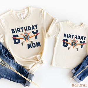 a pair of white t-shirts with the text "Birthday Boy Mom" printed on them, along with an illustration of an airplane. The t-shirts are laid out on a white background, and there are also some jeans visible in the image.