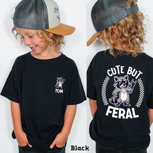 A young boy wearing a black t-shirt with a graphic of a raccoon and the text "CUTE BUT FERAL" printed on the back.
