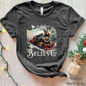 May include: Heather dark gray t-shirt featuring a detailed illustration of a vintage train with the word "BELIEVE" in a decorative font. The train is decorated for the holidays, with a snowy winter scene.