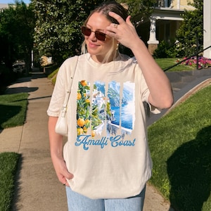 Puede incluir: Una camiseta de manga corta color crema con un estampado gráfico de la Costa Amalfitana con naranjos, edificios costeros y agua azul. El texto Amalfi Coast está impreso en letras azules debajo de la ilustración.