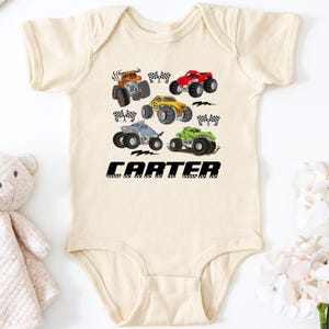 A baby onesie with a graphic of various monster trucks printed on it, along with the name "CARTER" printed below the trucks.