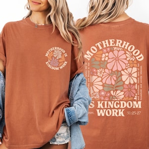 two women wearing orange t-shirts with floral designs. The t-shirts have text on the back that reads "Motherhood is kingdom work" and "31.25.27".