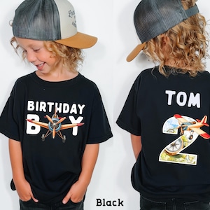 A young boy wearing a black t-shirt with a graphic of an airplane and the text "Birthday Boy" printed on it. The boy is also wearing a baseball cap and a black t-shirt with the number "2" printed on it.