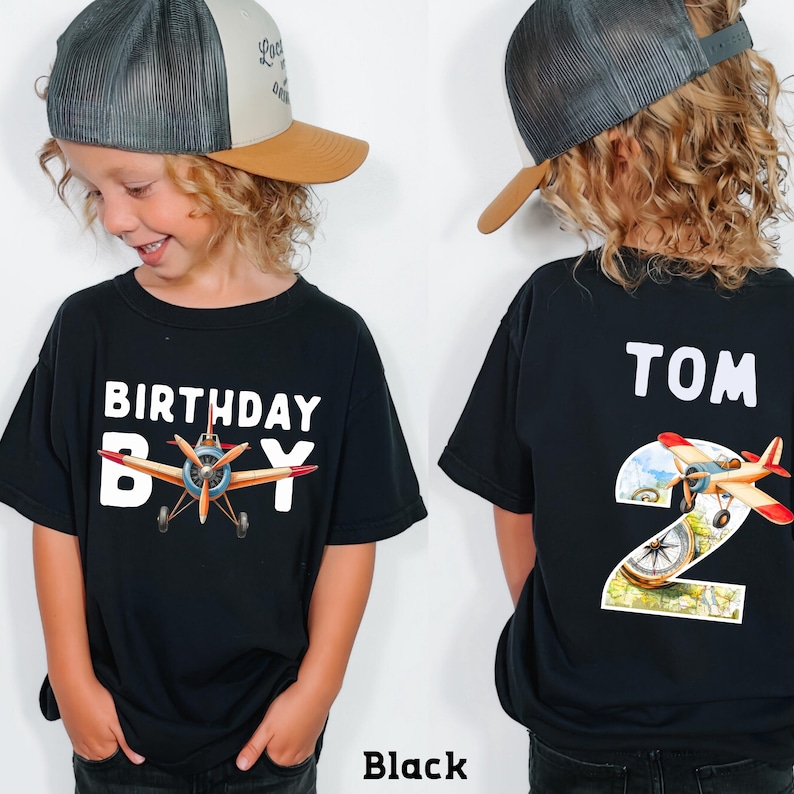 A young boy wearing a black t-shirt with a graphic of an airplane and the text "Birthday Boy" printed on it. The boy is also wearing a baseball cap and a black t-shirt with the number "2" printed on it.