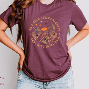 Hand-drawn graphic t-shirt featuring a dreamy desert landscape at sunset with surreal elements like planets in the sky, cacti, and animal skulls. The design includes the iconic lyric “On a Dark Desert Highway, Cool Wind in My Hair” from the classic Eagles song Hotel California. A perfect blend of retro, vintage, and boho aesthetics for music lovers and free spirits.