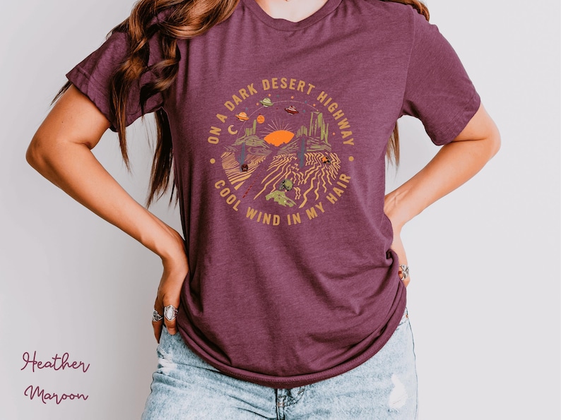 Hand-drawn graphic t-shirt featuring a dreamy desert landscape at sunset with surreal elements like planets in the sky, cacti, and animal skulls. The design includes the iconic lyric “On a Dark Desert Highway, Cool Wind in My Hair” from the classic Eagles song Hotel California. A perfect blend of retro, vintage, and boho aesthetics for music lovers and free spirits.