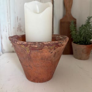 Könnte beinhalten: Eine weiße, batteriebetriebene Kerze steht in einem rustikalen Topf in Terrakotta-Farbe. Der Topf hat eine ausgestellte Form und ein abgenutztes Finish. Ein hölzernes Butterrührwerk und eine kleine Topfpflanze sind im Hintergrund.