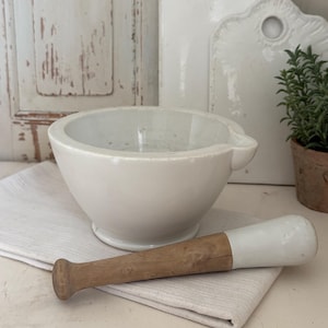 Könnte beinhalten: Ein weißes Keramik-Mörser- und Stößel-Set mit Holzgriff und weißem Ende. Zwei weiße Schneidebretter und eine kleine Topfpflanze befinden sich im Hintergrund. Mörser und Stößel liegen auf einem weißen Leinentuch.