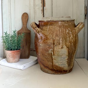 Könnte beinhalten: Ein großes, antikes braunes Keramikgefäß mit Griffen und Deckel. Daneben ein kleiner Terrakottatopf mit einer grünen Pflanze, ein hölzernes Butterbrett und ein gefaltetes weißes Tuch. Der Hintergrund ist eine verwitterte weiße Holztür.