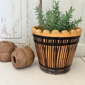 Könnte beinhalten: Ein Keramik-Blumentopf mit gewelltem Rand, verziert mit einem Muster aus vertikalen braunen und beigefarbenen Streifen sowie einem dunkelbraunen Boden. Eine grüne Pflanze steht im Topf, und zwei Knäuel braunes Garn liegen links daneben.