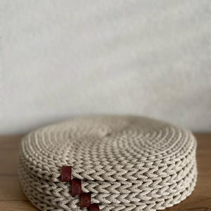 May include: A stack of beige, circular woven placemats. The placemats are made of a thick, textured material and have a braided appearance. Small, rectangular, dark red leather tags are attached to the side of the stack. The placemats are on a wooden surface.