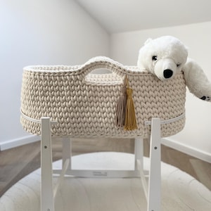 Pode incluir: Um berço de bebê de malha creme, com um suporte de balanço de madeira branca. O berço tem uma alça e borlas decorativas. Um urso de pelúcia branco está dentro. Projetado para recém-nascidos.