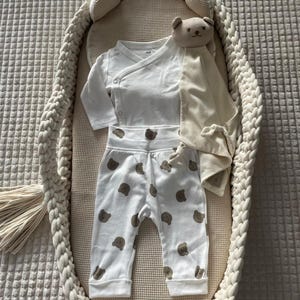May include: A baby clothing set featuring a white wrap-style long-sleeve shirt and white pants with brown bear prints. A beige security blanket with a bear head and a woven bassinet are also in the image.