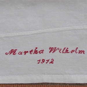 May include: White fabric with red cross-stitch embroidery. The embroidery reads "Martha Wilhelm" above the year "1912". The fabric has a seam and a stitched border.
