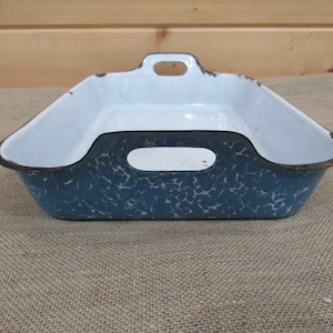 May include: A rectangular enamel baking dish featuring a blue and white marbled pattern on the exterior and a solid white interior. The dish includes two integrated handles on the shorter sides and shows signs of wear along the dark rim.