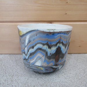May include: A cylindrical ceramic planter featuring a marbled pattern with swirling bands of blue, black, white, and tan colors. The interior of the vessel is finished in a smooth, solid white glaze, while the exterior displays a glossy finish.