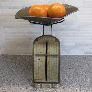 May include: An antique Imperial Computing Confectionery Scale with three oranges in the tray. The scale is made of metal and has a vintage look. The oranges are bright orange and sit on a gray tray.