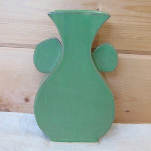 May include: A flat, green ceramic vase with a wide, rounded body, a narrow neck, and a flared rim. The vase features two symmetrical, rounded handles extending from the sides of the neck, and it has a smooth, solid green glazed finish.