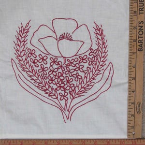 May include: A white fabric square featuring a red embroidered floral design of a large central flower surrounded by smaller blossoms and wheat-like stalks. A wooden ruler is placed along the right and bottom edges, showing a scale in inches.