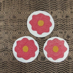 May include: Three circular coasters, each featuring a white base with a scalloped-edge pink flower shape appliqued in the center. Each pink flower has a solid yellow circular center. The coasters are arranged in a triangular formation.