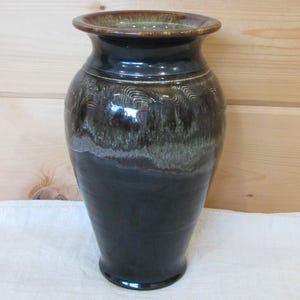 May include: A ceramic vase with a glossy finish, featuring a dark black base that transitions into a mottled brown and cream glaze near the wide, flared rim. The upper body displays subtle, carved horizontal wave patterns in the clay.