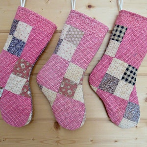 May include: Three handmade Christmas stockings. Each stocking is primarily pink with a patchwork design of various patterned fabrics in cream, brown, and navy blue. The stockings are hanging against a light wood background.