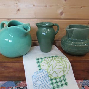 May include: Three ceramic pitchers in varying shades of green. The left pitcher is a round, light mint green. The center pitcher is a smooth, dark olive green. The right pitcher is a sage green with horizontal ribbed detailing around the base.