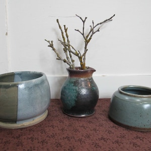 May include: Three ceramic vessels: a round bowl with a split olive and light blue glaze, a small dark pitcher with a teal and brown mottled finish containing bare twigs, and a low, wide, speckled light blue bowl with a brown rim.