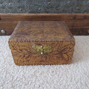 May include: A small rectangular wooden box featuring a dark brown pyrography floral pattern on the lid and front panels. The box is secured with a central brass-toned metal latch and has a smooth, light brown finish across its exterior surfaces.
