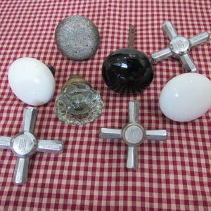May include: A collection of seven assorted hardware knobs and faucet handles. Included are two smooth white oval knobs, one textured grey round knob, one clear faceted glass knob, one black round knob, and two silver metal cross-shaped faucet handles.