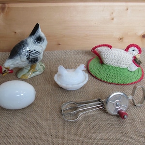 May include: A collection of kitchen-themed items, including a ceramic chicken figurine, a white egg, a white egg-shaped dish, a crocheted chicken trivet, and a vintage egg beater. The items are arranged on a burlap surface.