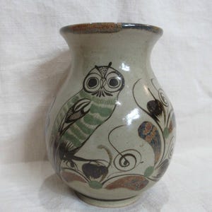May include: A light gray ceramic vase with a flared rim, featuring a hand-painted illustration of an owl in green and black tones, surrounded by decorative floral and swirling leaf motifs in shades of brown, orange, and green.