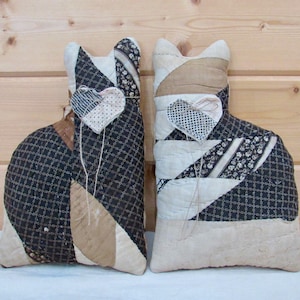 May include: Two cat-shaped stuffed fabric pillows featuring patchwork designs in black, white, and tan. Each pillow has a small, heart-shaped fabric ornament attached with a thin string. The patterns include geometric grids, floral prints, and solid colors.