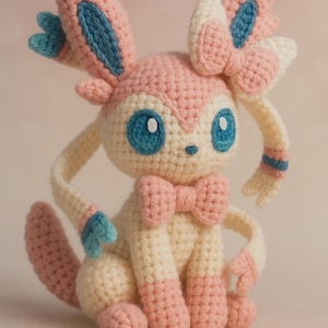 Op de afbeelding: Een handgemaakt gehaakt Sylveon-knuffel. Het speelgoed heeft voornamelijk roze en crèmekleurige kleuren met blauwe accenten. Het heeft grote blauwe ogen, een strik en een strik op zijn hoofd. De knuffel zit.