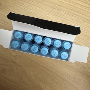 Op de afbeelding: Een witte rechthoekige doos met elf cilindervormige voorwerpen met lichtblauwe doppen. Op elke dop staat het nummer 822 in een donkerblauw lettertype gedrukt. De doos is aan de bovenkant open met een zwarte binnenflap aan de rand.