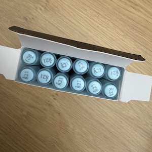Puede incluir: Una caja de cartón blanca que contiene diez artículos cilíndricos dispuestos en dos filas. Cada artículo tiene una tapa azul claro con el número 821 impreso en una fuente oscura. Los artículos están colocados verticalmente en el envase.