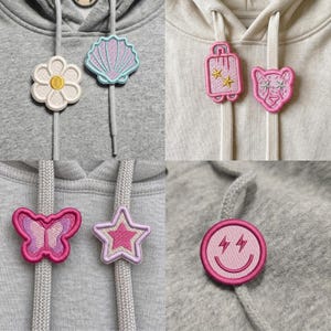 May include: A collection of embroidered hoodie drawstring charms featuring various shapes including a white flower, a light blue seashell, a pink suitcase with stars, a pink leopard head, a pink butterfly, a pink star, and a pink smiley face.