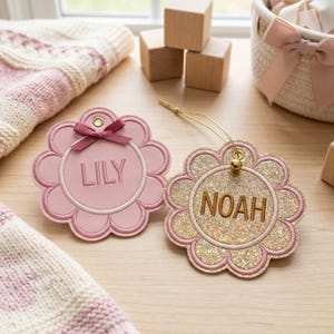 May include: Two flower-shaped ornaments with scalloped edges. One is matte pink with the name LILY embroidered in pink and a small pink ribbon bow. The other is gold glitter with the name NOAH embroidered in brown and a gold hanging string.