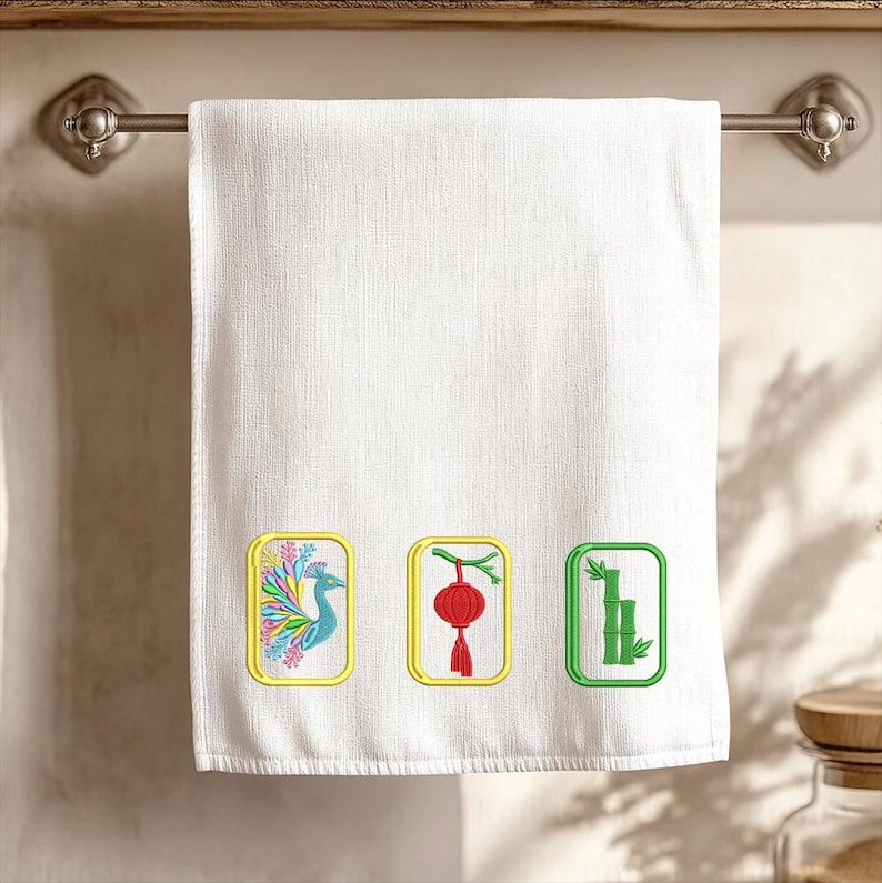 Puede incluir: Una toalla de mano blanca con tres parches rectangulares bordados. El parche izquierdo muestra un pavo real colorido, el central una linterna roja y el derecho tallos de bamb&uacute; verdes, todos dispuestos horizontalmente.