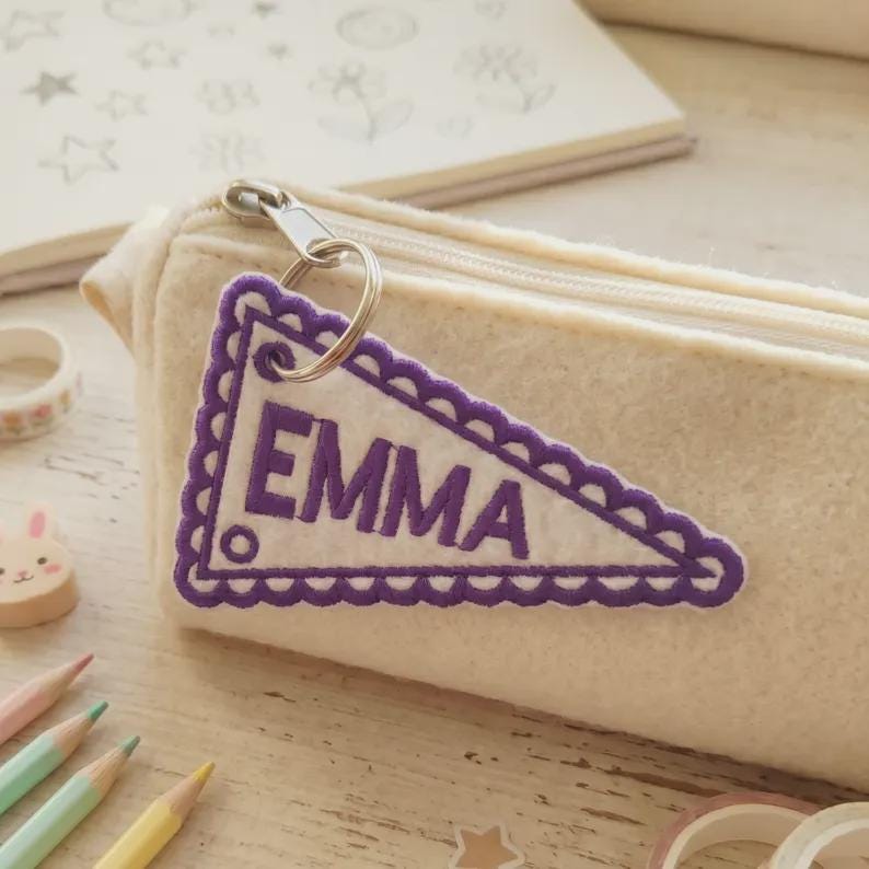 May include: A cream-colored pencil case with a zipper, featuring a purple pennant-shaped keychain. The keychain has the name "EMMA" embroidered in purple. The background includes pencils and a notebook.