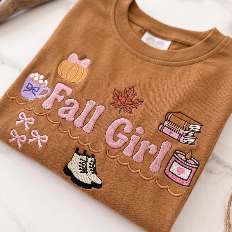 May include: Brown crew neck t-shirt with embroidered fall-themed graphics. Features a pumpkin, maple leaf, books, candle, and the text "Fall Girl" in pink. Includes a mug of hot chocolate and a pair of boots.