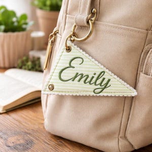 May include: A beige bag with gold-colored hardware and a triangular tag. The tag is light green with a striped pattern and the name "Emily" embroidered in green. The tag is attached to the bag with a gold ring.