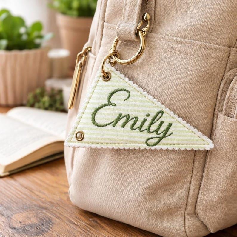 May include: A beige bag with gold-colored hardware and a triangular tag. The tag is light green with a striped pattern and the name "Emily" embroidered in green. The tag is attached to the bag with a gold ring.