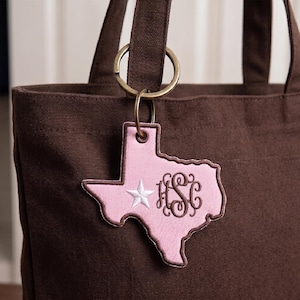 May include: A pink Texas-shaped keychain with a white star and monogram, attached to a brown tote bag with a gold ring. The keychain has a brown border and is hanging from a brown tote bag.