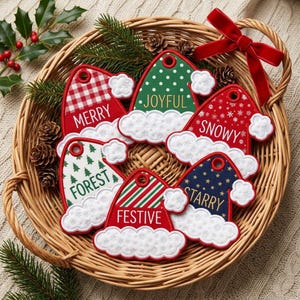 May include: A collection of six felt Santa hat ornaments in a wicker basket. Each hat features a unique pattern and text: MERRY in red plaid, JOYFUL in green polka dot, SNOWY in red snowflake, FOREST in white tree print, FESTIVE in red stripes, and STARRY in navy blue stars.