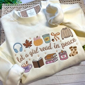 May include: Cream-colored sweatshirt with embroidered fall-themed graphics and the phrase "let a girl read in peace." The design includes a pumpkin, leaf, books, headphones, and a cup of coffee.