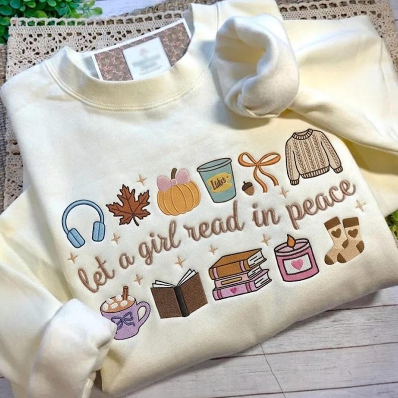 May include: Cream-colored sweatshirt with embroidered fall-themed graphics and the phrase "let a girl read in peace." The design includes a pumpkin, leaf, books, headphones, and a cup of coffee.