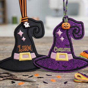 May include: Two witch hat ornaments featuring embroidered details. The black hat has the name Liam, a ghost charm, and orange-striped ribbon. The purple glitter hat has the name Charlotte, a pumpkin bead, and a green spiderweb-patterned ribbon.